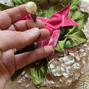 May include: A vintage hat pin with a white pearl-like head and a gold-toned metal base. The pin is held in a hand against a background of pink and green fabric flowers.