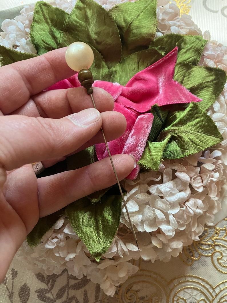 May include: A vintage hat pin with a white pearl-like head and a gold-toned metal base. The pin is held in a hand against a background of pink and green fabric flowers.