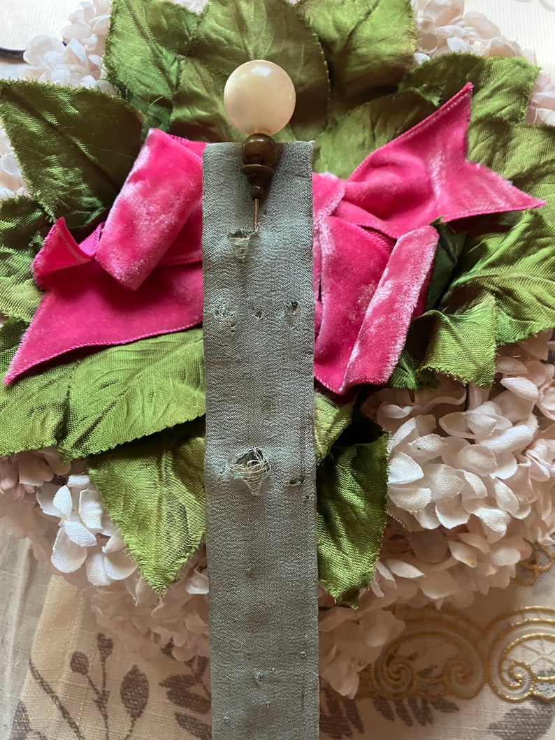May include: A white and brown pin with a pearl-like top. The pin is attached to a gray fabric ribbon with holes.