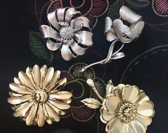 May include: Four gold and silver flower brooches. Two of the brooches are gold and have a daisy shape. One brooch is silver and has a bow shape. The other brooch is silver and has a more abstract flower shape.