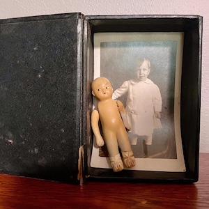 May include: A small, light-colored, jointed doll with painted facial features stands inside an open, black, rectangular box. Behind the doll is a black and white vintage photograph of a child wearing a light-colored dress, positioned vertically within the box.