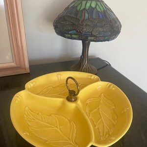 May include: A yellow, three-sectioned serving dish with leaf designs. The dish has a brass handle and is sitting on a dark surface. A Tiffany-style lamp with a stained glass shade is in the background.