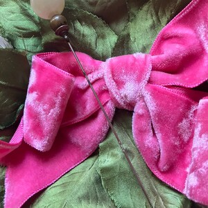 May include: A vintage hat pin with a white glass ball head and a silver metal pin. The pin is placed on a pink velvet bow.