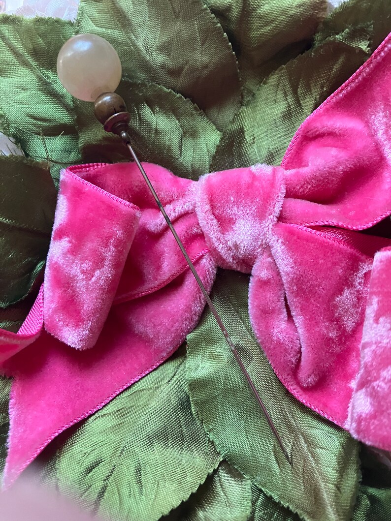 May include: A vintage hat pin with a white glass ball head and a silver metal pin. The pin is placed on a pink velvet bow.