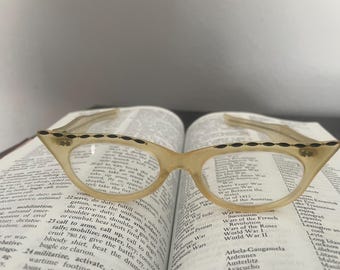 May include: Vintage cat-eye eyeglasses with a light beige frame and black decorative accents. The glasses rest on an open book with printed text. The frames have a classic, retro design.