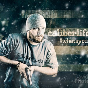 May include: A man wearing a gray beanie and a gray t-shirt with the text "Caliber Life" printed on it. He is holding a gun in his right hand. The background is a blurred image of an American flag with snow falling.
