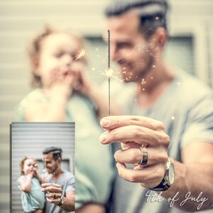 May include: A father and daughter are holding sparklers and smiling. The father is wearing a gray shirt and the daughter is wearing a light blue shirt. The text "4th of July" is written in white cursive on the bottom right of the image.