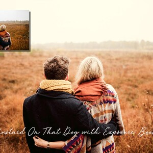 May include: A couple wearing scarves and jackets stand in a field of tall grass, looking out at the horizon. The grass is a mix of brown and green, and the sky is a light blue.