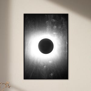 May include: A black and white abstract art print featuring a black circle with a white halo, resembling a solar eclipse. The background is a dark gray with a textured, brushstroke effect.