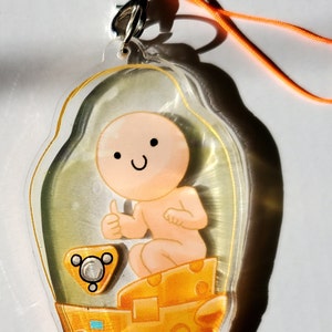 Puede incluir: Un llavero de plástico transparente con un personaje de dibujos animados sonriente y con el pulgar hacia arriba, sentado en un objeto amarillo con forma de queso. El personaje está dentro de una cúpula de plástico transparente.