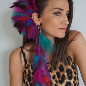 Può includere: Un'orecchino a piuma vibrante nei toni del viola, verde acqua e rosso. L'accessorio è attaccato all'orecchio e scende. La modella indossa un top con stampa leopardata.