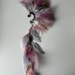 May include: A decorative ear cuff featuring a cascade of pink, gray, and brown feathers. The feathers are attached to a black wire frame with a spiral accent. The design is asymmetrical, with a cluster of feathers at the top and a longer, flowing section below.
