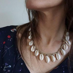 Peut inclure: Un collier de coquillages blancs avec un cordon brun. Le collier est composé de petites coquilles blanches enfilées sur un cordon brun. Le collier est porté par une personne aux longs cheveux bruns.
