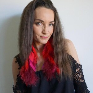 Pode incluir: Uma pessoa com cabelo castanho comprido usando um top preto com os ombros de fora e brincos de penas vibrantes. Os brincos apresentam penas vermelhas e rosa, adicionando uma explosão de cor contra o top escuro e o cabelo.