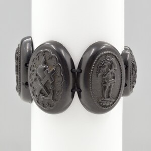 May include: A black jet bracelet with oval-shaped links featuring raised, carved designs. The links include a heart with a cross, an anchor, and a figure holding a tree.