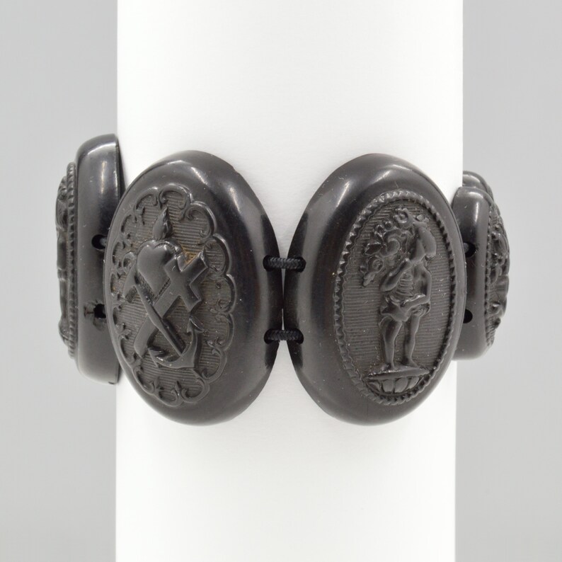 May include: A black jet bracelet with oval-shaped links featuring raised, carved designs. The links include a heart with a cross, an anchor, and a figure holding a tree.