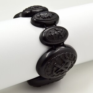 May include: Black jet bracelet with oval-shaped beads featuring intricate carved designs. Each bead has a different design, including a floral motif, a heart, and an anchor.