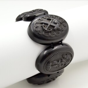 May include: A black jet bracelet with oval-shaped links featuring carved designs. The links include an anchor and cross, a cherub, and a floral motif.