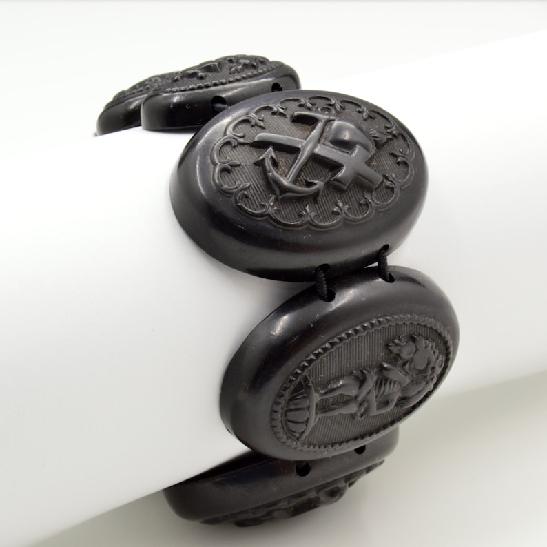 May include: A black jet bracelet with oval-shaped links featuring carved designs. The links include an anchor and cross, a cherub, and a floral motif.