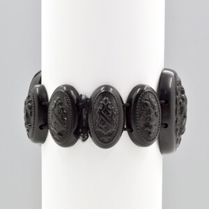 May include: A black jet bracelet with oval-shaped links featuring an embossed design. Each link is connected by a black elastic band.