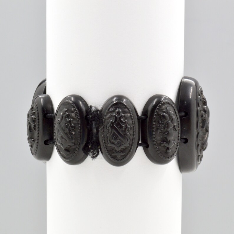May include: A black jet bracelet with oval-shaped links featuring an embossed design. Each link is connected by a black elastic band.
