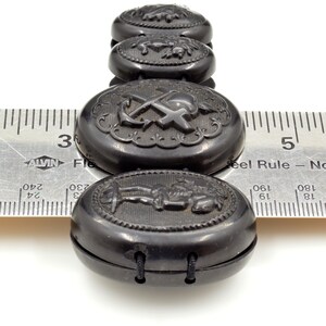 May include: A black jet button bracelet with oval-shaped buttons featuring various engraved designs, including an anchor and cross, a figure, and a floral motif. The bracelet is measured against a ruler.