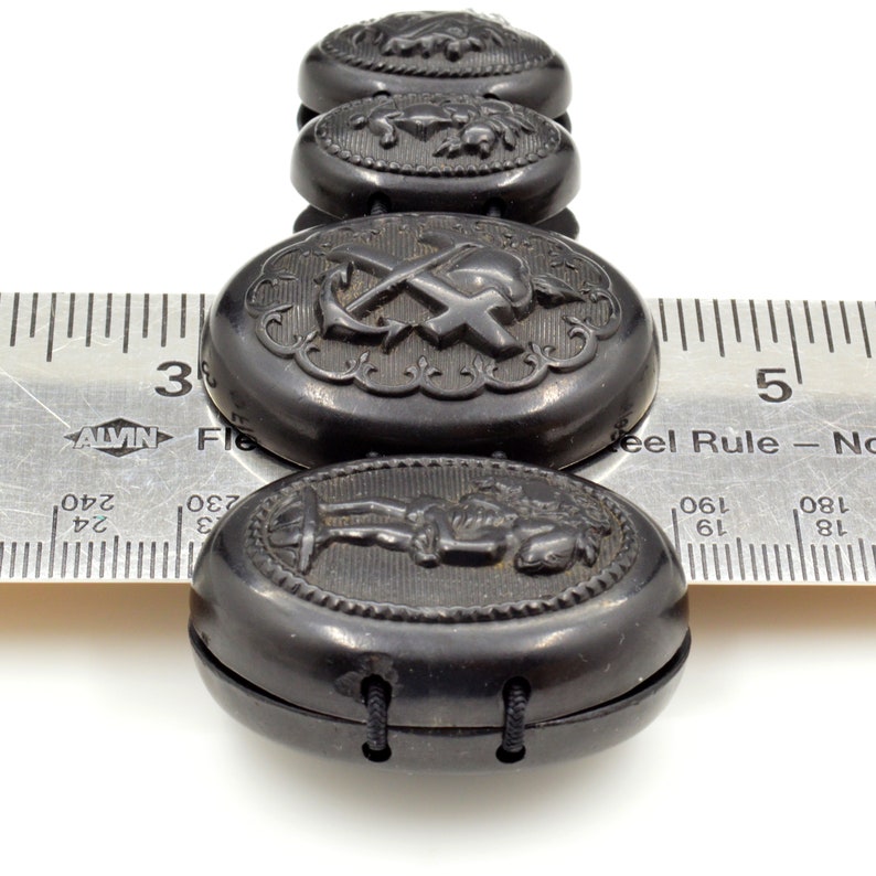May include: A black jet button bracelet with oval-shaped buttons featuring various engraved designs, including an anchor and cross, a figure, and a floral motif. The bracelet is measured against a ruler.