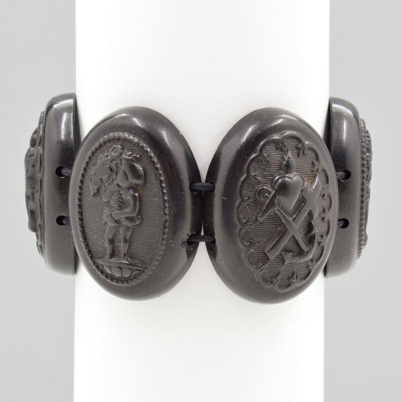 May include: A black jet bracelet with five oval-shaped links. Each link features a different carved design, including a cherub, a heart, an anchor, and two floral designs.