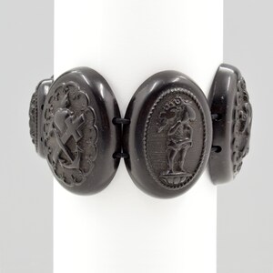 May include: A black jet bracelet with oval-shaped links. Each link features a different carved design, including a heart, cross, anchor, and a figure holding a flower.