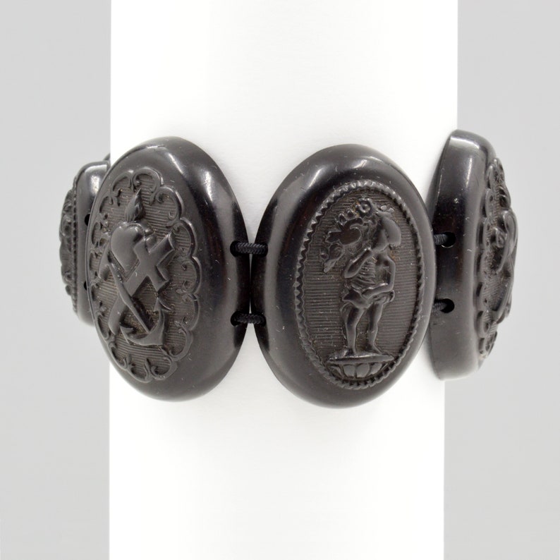 May include: A black jet bracelet with oval-shaped links. Each link features a different carved design, including a heart, cross, anchor, and a figure holding a flower.
