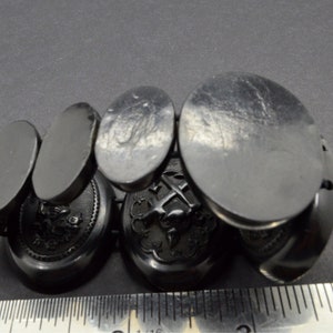 May include: A set of six black oval cufflinks with intricate engraved designs. The cufflinks are made of a smooth, polished material and feature a variety of designs, including a cross, a bird, and a floral motif.