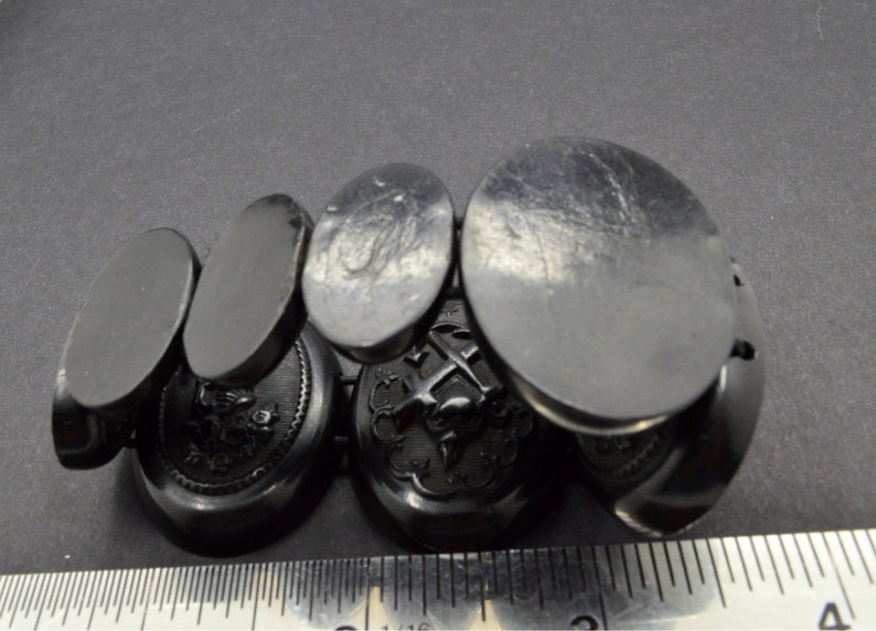 May include: A set of six black oval cufflinks with intricate engraved designs. The cufflinks are made of a smooth, polished material and feature a variety of designs, including a cross, a bird, and a floral motif.