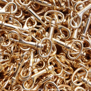 Puede incluir: Una imagen de primer plano de una pila de cierres giratorios de metal dorado. Los cierres son todos del mismo tamaño y forma, y están dispuestos en un patrón aleatorio.
