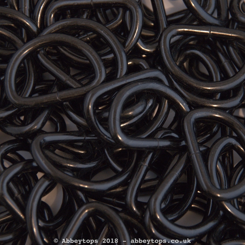 May include: A close-up image of a pile of black metal D-rings. The rings are all the same size and shape, and they are arranged in a random pattern.