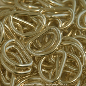 May include: A close-up of a pile of gold-colored metal D-rings. The rings are all the same size and shape, and they are arranged in a random pattern.