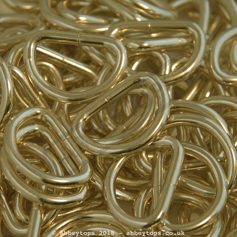 May include: A close-up of a pile of gold-colored metal D-rings. The rings are all the same size and shape, and they are arranged in a random pattern.