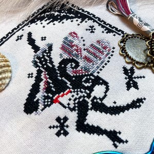 May include: A cross-stitch embroidery on light-colored fabric featuring a black silhouette of a winged creature with red and variegated thread accents. The design includes small star motifs, a dragonfly, and a decorative border. Embroidery floss and scissors are visible.
