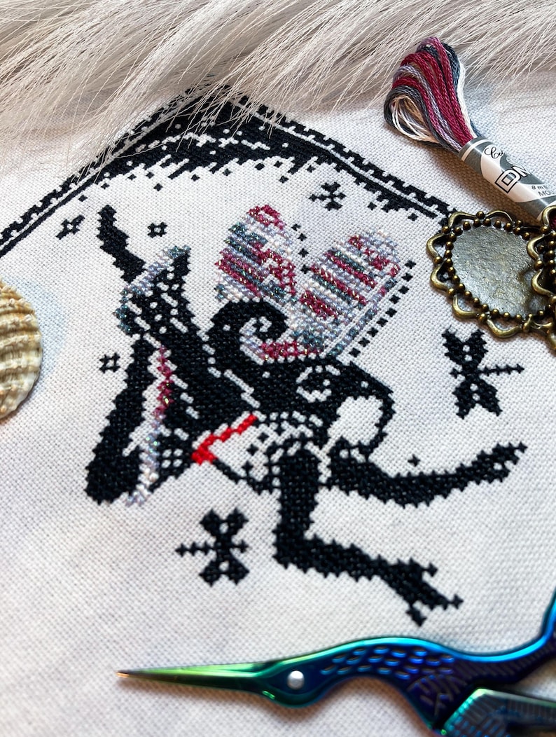 May include: A cross-stitch embroidery on light-colored fabric featuring a black silhouette of a winged creature with red and variegated thread accents. The design includes small star motifs, a dragonfly, and a decorative border. Embroidery floss and scissors are visible.