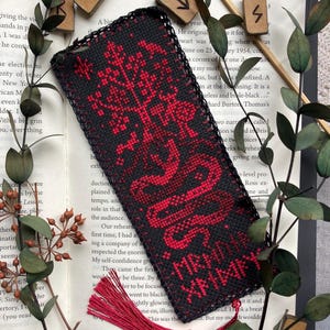 K&ouml;nnte beinhalten: Schwarz-rotes besticktes Lesezeichen mit Quaste. Das Lesezeichen zeigt ein rotes Baum- und Schlangenmuster mit Text. Das Lesezeichen liegt auf einem offenen Buch mit Gr&uuml;n und Holzrunen.