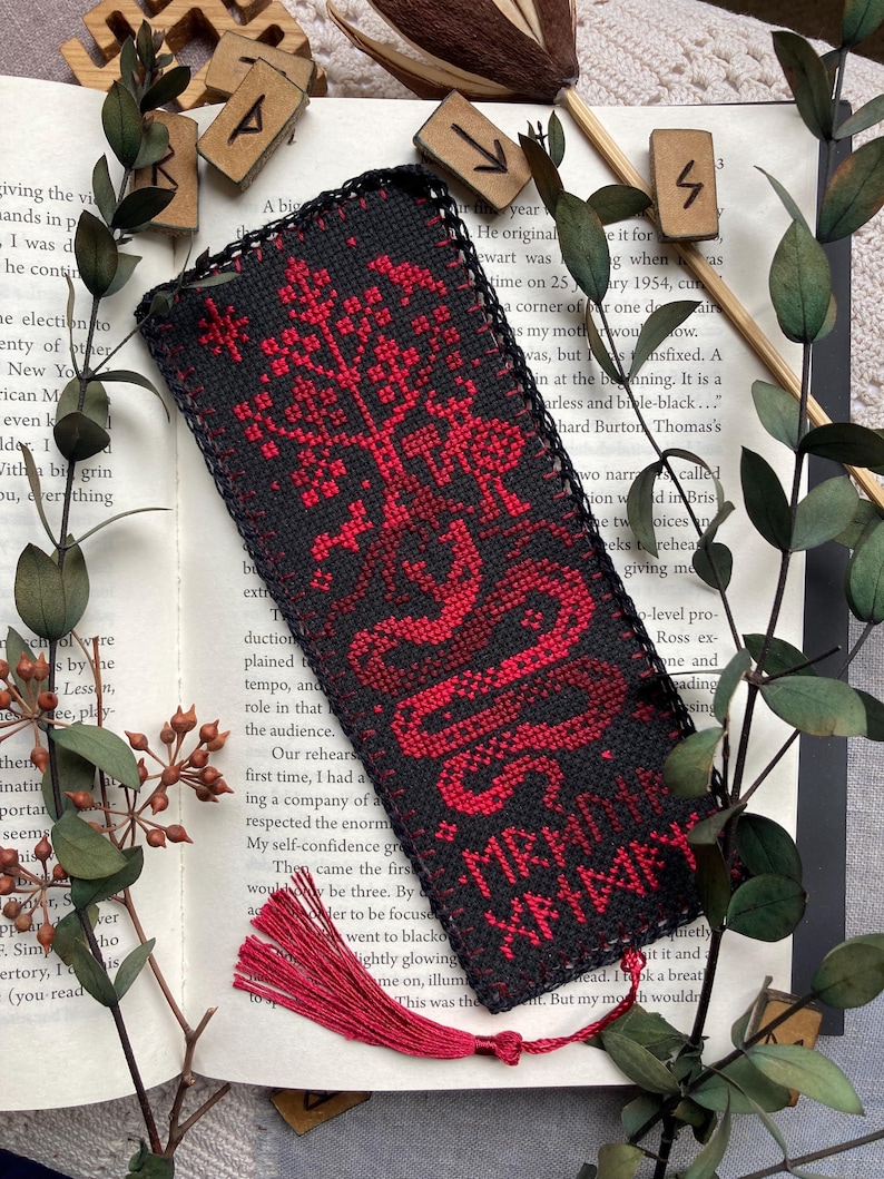 K&ouml;nnte beinhalten: Schwarz-rotes besticktes Lesezeichen mit Quaste. Das Lesezeichen zeigt ein rotes Baum- und Schlangenmuster mit Text. Das Lesezeichen liegt auf einem offenen Buch mit Gr&uuml;n und Holzrunen.