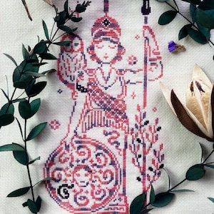 May include: Cross-stitch embroidery of a figure in red, pink, and black, holding a staff and an owl. The figure is adorned with a patterned headdress and clothing. The background is a light fabric, with green leaves and dried flowers framing the image.