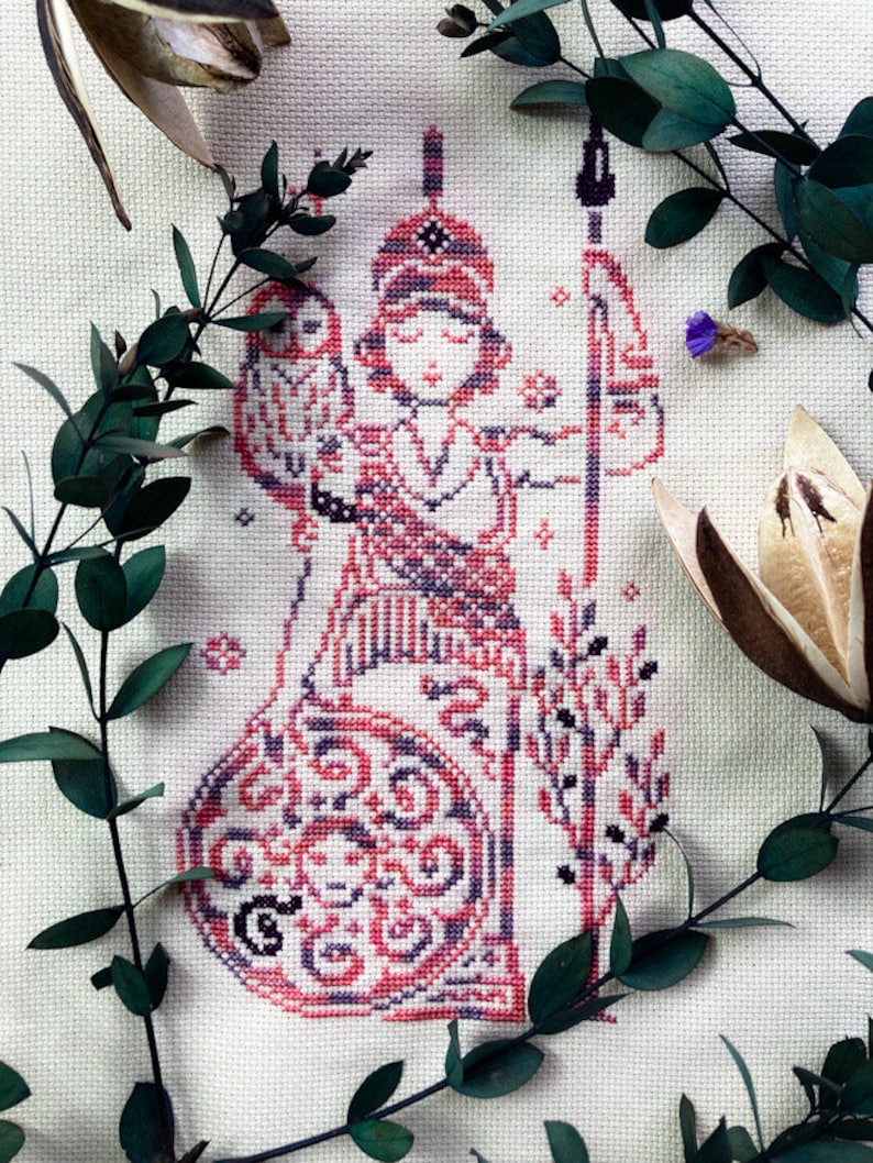 May include: Cross-stitch embroidery of a figure in red, pink, and black, holding a staff and an owl. The figure is adorned with a patterned headdress and clothing. The background is a light fabric, with green leaves and dried flowers framing the image.