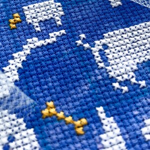 May include: Close-up of a cross-stitch embroidery piece with a blue background. White thread forms a building and stars, with gold thread accents. The texture of the fabric is visible.