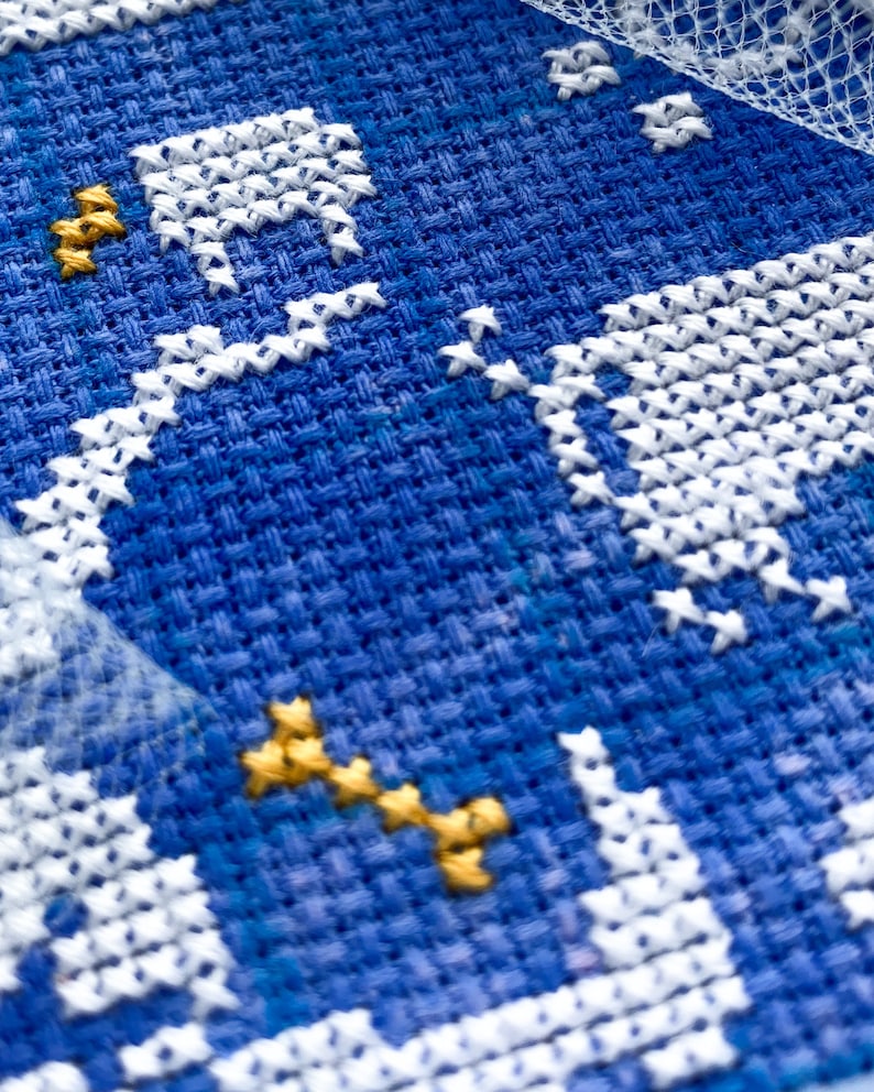 May include: Close-up of a cross-stitch embroidery piece with a blue background. White thread forms a building and stars, with gold thread accents. The texture of the fabric is visible.