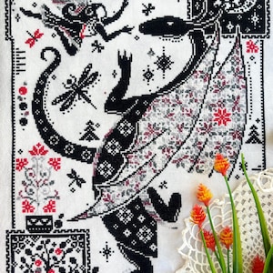 May include: A cross-stitch embroidery on white fabric featuring a large black dragon with patterned wings, a small fairy, dragonflies, and floral motifs. The design uses a black, white, and red color palette with intricate geometric borders.