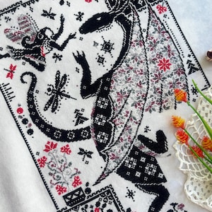 May include: A cross-stitch embroidery on white fabric featuring a large black dragon with patterned wings, a small fairy, dragonflies, and floral motifs in black, red, and grey thread. The design includes geometric borders and intricate details.