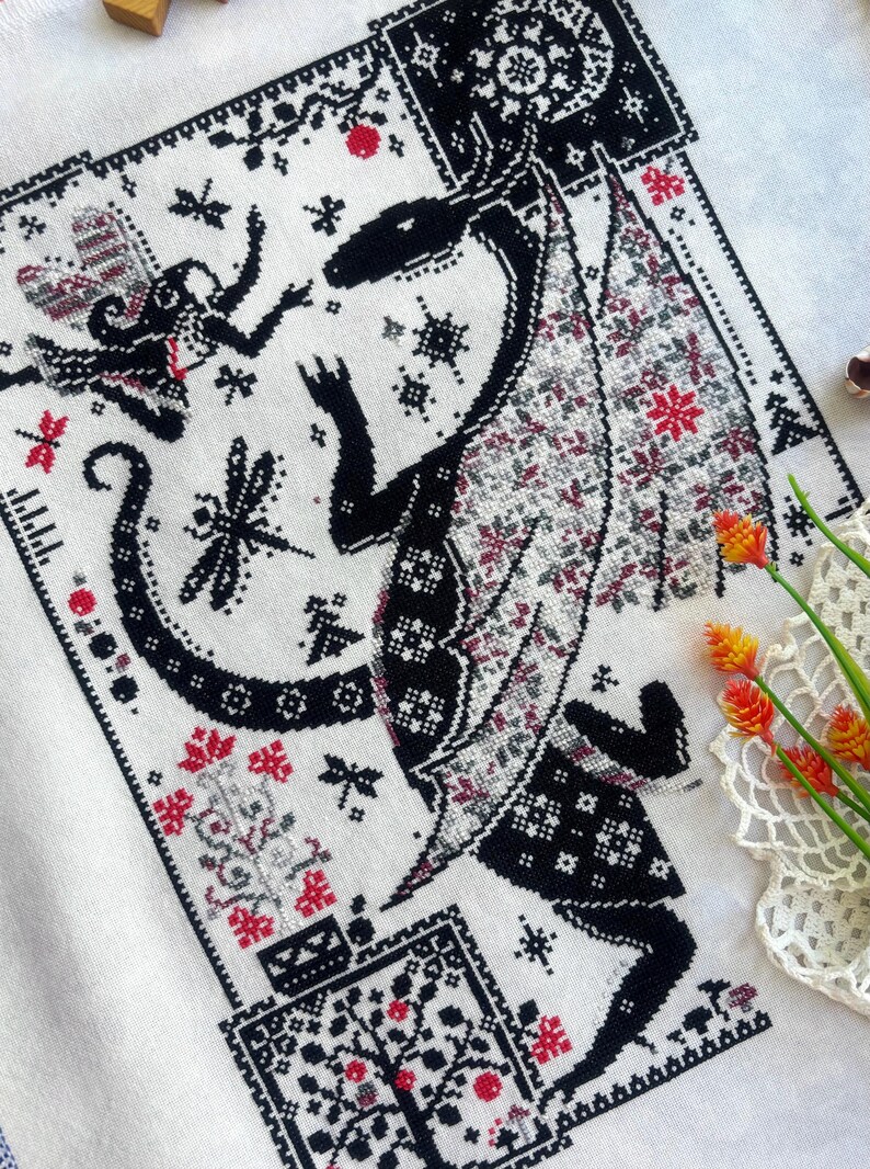 May include: A cross-stitch embroidery on white fabric featuring a large black dragon with patterned wings, a small fairy, dragonflies, and floral motifs in black, red, and grey thread. The design includes geometric borders and intricate details.
