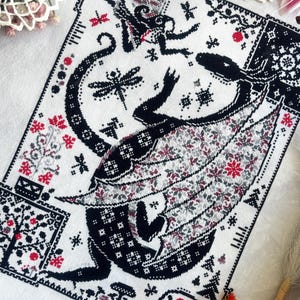 May include: A cross-stitch embroidery on white fabric featuring a large black dragon with patterned wings, a smaller winged creature, dragonflies, mushrooms, and geometric borders. The design uses black, white, and red thread to create intricate details.