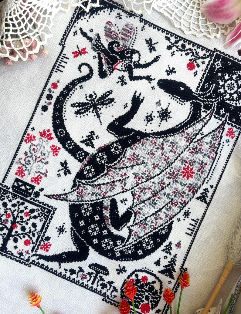 May include: A cross-stitch embroidery on white fabric featuring a large black dragon with patterned wings, a smaller winged creature, dragonflies, mushrooms, and geometric borders. The design uses black, white, and red thread to create intricate details.
