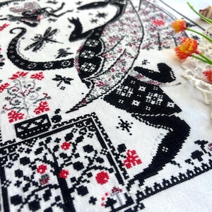 May include: A white fabric piece featuring a detailed cross-stitch embroidery pattern in black and red thread. The design depicts a large winged creature, a dragonfly, and floral motifs arranged within a decorative border on the textile.